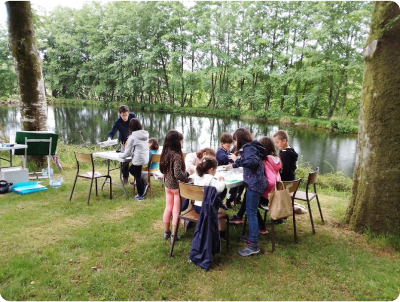 Photo prise lors d'un atelier animé par le plasticien Pierre Quentel, dans cette photo on voit des enfants autour d'une table entrain de faire de la peinture et au fond on peut voir le peinture entrain de les aider