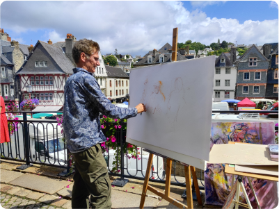 Photo prise pour la galerie du plasticien pierre Quentel, dans cette photo on voit Pierre entrain de réaliser une toile de peinture en direct devant la galerie Des Fous, des Sages à Morlaix et derrière lui on voit le marché de samedi à Morlaix