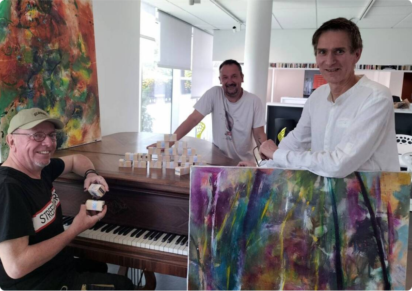 Phot prise pour la page la presse en parle de l' artiste breton Pierre Quentel lors de l'expo au Roudour avec l'artiste Dominic, on voit pierre derrière une de ses toiles, et les collaborateurs un derrière le piano et l'autre assis