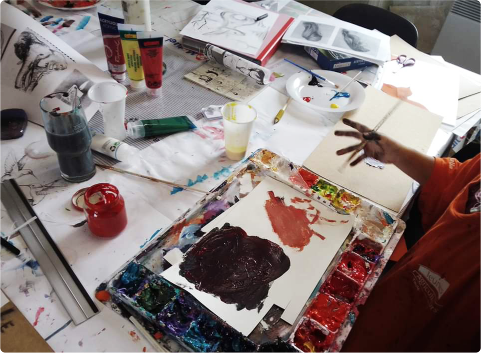 Photo prise pour la page d'accueil de Pierre Quentel, dans cette photo on voit une table pleine des outils de dessins tel que une palette, des tubes de peinture et des papiers ainsi que la main d'un enfant pleine de peinture